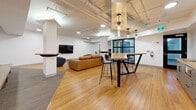 Located in Toronto, ON, this single-floor amenity space offers a generous, open-concept layout designed to accommodate a variety of social and recreational activities. The interior showcases an industrial-inspired aesthetic, with exposed piping and ductwork running along the white-painted ceiling, complemented by a combination of warm hardwood flooring and multi-tone carpet tiles that subtly define different functional zones.

The main lounge area features a tan leather sofa and a plush blue accent chair arranged around a low coffee table, with a wall-mounted flat-screen television nearby. A structural column with integrated ledge shelving adds architectural character while dividing the space thoughtfully. Adjacent to the lounge, a high-top communal table with black bar stools sits beneath a cluster of pendant lights, creating an ideal spot for casual gatherings or collaborative work.

The kitchen area is anchored by a white subway tile backsplash, a stainless steel refrigerator, and a bar-height counter with seating, making it well-equipped for entertaining. A dedicated recreation zone includes a foosball table set on carpeted flooring, with a lush fiddle-leaf fig plant adding a touch of greenery. A wall-mounted split AC unit ensures year-round comfort. The space is completed by bold black doors and curated abstract wall art, lending a polished, contemporary feel throughout.