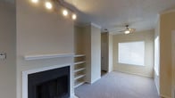 This single-floor space is designed with comfort and practicality in mind, offering a well-appointed living area that balances functional features with a warm, inviting ambiance. The main living area is anchored by a classic white-mantled fireplace, which serves as a natural focal point of the room. Flanking the fireplace is a set of built-in open shelving units, providing ample display and storage space while adding architectural character to the wall. The living area is finished with plush wall-to-wall carpeting that adds warmth underfoot, and a ceiling fan overhead ensures year-round comfort and air circulation. A track lighting fixture mounted near the fireplace wall provides targeted illumination, complementing the natural light that filters in through a window dressed with horizontal blinds. The textured ceiling adds depth and dimension to the space. A hallway visible from the living area leads further into the home, hinting at additional rooms beyond. The overall color palette is neutral and understated, with soft beige walls and white trim creating a clean, versatile backdrop well-suited to a variety of interior styles. This space offers a functional and comfortable layout ideal for everyday living.