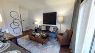 This single-story space showcases a thoughtfully designed living area that blends contemporary style with inviting warmth. The room features rich, dark hardwood flooring that anchors the space and complements the carefully curated furnishings throughout. A plush charcoal-blue sofa serves as the focal point, dressed with an assortment of patterned and solid throw pillows in navy, gold, and teal tones. A cognac leather armchair with nailhead trim adds a classic touch alongside it.

A substantial wood-slatted coffee table with dark metal legs sits atop a light-toned, abstract area rug, offering both style and function. Decorative accents, including a geometric gold orb and textured vessels, add visual interest to the tabletop. The walls are painted in a clean, neutral white, allowing a bold black geometric wall art piece to stand out prominently above the sofa.

Ambient lighting is provided by two coordinating table and floor lamps, creating a warm and welcoming atmosphere. A white paneled entry door is visible to the left, alongside a floor-level vent, indicating practical, well-integrated utility features. The overall ambiance is modern, cohesive, and polished.
