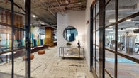 This single-floor space delivers a compelling blend of industrial and contemporary design. Exposed concrete ceilings with visible ductwork and structural beams set an urban, loft-like tone throughout. Steel-framed glass doors and partition walls are a defining architectural feature, dividing zones while maintaining visual openness and allowing natural light to flow freely across the interior.

The main lounge area is generously proportioned, furnished with an eclectic mix of colorful modern seating—including bold accent chairs and plush sofas—arranged atop textured area rugs over polished concrete floors. A wall-mounted display and built-in wood shelving anchor one end of the room, while large grid-paned windows frame greenery views outside.

A well-appointed kitchen and social hub features a central island with bar seating, pendant lighting, stainless steel appliances, tiled backsplash, and a mounted television. A cozy booth-style seating nook with teal upholstery and a dark textured accent wall offers a more intimate setting.

Private meeting areas enclosed by floor-to-ceiling steel-and-glass doors provide focused workspaces with tree-lined outdoor views. Throughout, the design balances raw industrial elements with curated furnishings, vibrant artwork, and warm wood accents, creating a dynamic and functional environment.