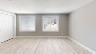 This single-floor space presents a clean, modern aesthetic throughout, characterized by neutral gray walls, white trim, and light-toned vinyl plank flooring that flows consistently across all areas. The textured ceilings complement the understated palette, lending a cohesive feel to the interior.

The living area benefits from multiple windows fitted with horizontal blinds, allowing ample natural light to fill the space. A ceiling fan adds both functionality and a casual design element near the entry door, which features frosted glass panel accents.

The kitchen is efficiently designed with a peninsula layout that partially separates it from the main living area. Gray shaker-style cabinetry is paired with light countertops and a classic white subway tile backsplash, creating a crisp, contemporary look. Stainless steel appliances — including a range, over-the-range microwave, and refrigerator — complete the kitchen's practical setup. A stainless steel sink sits beneath the upper cabinets, maximizing counter workspace.

Additional rooms accessible via a central hallway maintain the same consistent flooring and neutral color scheme. Recessed lighting and ceiling-mounted fixtures provide well-distributed illumination throughout. Overall, the space offers a bright, move-in-ready environment with a cohesive modern finish.