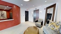 This single-floor space presents a polished and contemporary interior designed to make a strong first impression. A bold, curved terracotta-red accent wall commands attention upon entry, adding warmth and a sculptural architectural element to the otherwise crisp white surroundings. Recessed lighting throughout the ceiling ensures the space is well-lit, contributing to its clean and professional ambiance.

The area is appointed with modern furnishings, including a sleek leather lounge chair with a chrome base and a rounded gold ottoman coffee table that adds a luxurious, metallic accent to the space. A plush sofa is also visible, offering comfortable seating for visitors or waiting guests.

A stainless steel elevator with a polished finish serves as a central functional feature, accompanied by a standard access door marked with an illuminated exit sign, indicating this space functions as a shared common area or lobby within a larger building. Wall-mounted control panels and call buttons are neatly integrated into the white walls.

The flooring appears to be a light-toned, smooth surface that complements the overall neutral palette. Together, the architectural details and carefully selected furnishings create a sophisticated, welcoming environment that balances functionality with contemporary design aesthetics.