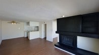 This single-floor space offers a clean, open layout that seamlessly connects the living area to the kitchen. The living room is anchored by a striking floor-to-ceiling black fireplace surround, complete with a tiled hearth and dark mantel, serving as a bold focal point against the neutral white walls. Hardwood-style flooring runs throughout, adding warmth and continuity to the space. The textured popcorn ceiling provides a classic finish overhead. The kitchen is efficiently designed with white cabinetry, a peninsula countertop that opens toward the living area, and essential appliances including a refrigerator and sink. The open pass-through design between the kitchen and living space enhances the sense of openness and allows for easy interaction across both areas. A hallway visible from the main living space suggests access to additional rooms beyond. The overall aesthetic is straightforward and functional, with a neutral color palette of white and black creating a high-contrast, easy-to-personalize environment. The space is currently vacant, allowing prospective occupants to clearly envision furniture placement and interior customization.