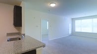 This single-floor space offers a clean, open layout designed for comfortable everyday living. The main living area is generously proportioned, featuring soft wall-to-wall carpeting in a neutral tone that complements the crisp white walls throughout. Flush-mount ceiling lighting provides consistent illumination, while multiple windows allow natural light to filter into the space, creating a bright and welcoming atmosphere.

A partial view of the kitchen reveals a dark-based counter topped with granite, adding a touch of refined detail to the functional space. The kitchen transitions naturally into the open living area, supporting an inclusive, connected floor plan.

Two doorways are visible from the main living space — one leading to a secondary room bathed in natural light from its own window, and another door that likely accesses an additional room or hallway. Both openings reinforce the sense of flow and connectivity throughout the unit.

White baseboards frame each room neatly, contributing to the clean aesthetic. Wall-mounted fixtures such as a thermostat and outlets are conveniently positioned. Overall, the space presents a practical, well-lit, and move-in-ready environment with a modest yet functional design.
