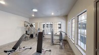 This fitness center offers a clean, functional space designed to support a well-rounded workout routine. The room is outfitted with an assortment of strength and resistance training equipment, including a cable machine, a multi-function weight station, a flat/incline bench, and a seated resistance machine, providing options for both upper and lower body exercises. The space is finished with neutral, light-toned walls and durable patterned carpet flooring suited for a fitness environment. Recessed lighting and a skylight keep the interior bright and evenly illuminated, while a row of large multi-pane windows along two walls invites abundant natural light and provides views to the surrounding exterior, enhancing the open feel of the space. The layout is thoughtfully arranged to allow ease of movement between equipment. Overall, the fitness center presents a practical and comfortable environment suitable for daily exercise, with a simple, no-frills aesthetic that keeps the focus on functionality and usability.