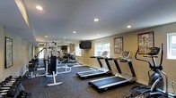 This fitness center offers a comprehensive workout environment designed to accommodate a variety of exercise routines. The space is finished with neutral-toned walls and recessed lighting overhead, creating a clean and functional atmosphere throughout. Carpeted flooring in a muted tone covers the workout area, providing comfort underfoot while defining the exercise zones.

The cardio section is prominently featured and includes multiple treadmills arranged in a row, as well as an elliptical machine and a stationary bike. A wall-mounted flat-screen television is positioned at the front of the room, visible from the cardio equipment, making it convenient for users during longer workout sessions. Natural light enters through several windows along the walls, contributing to an open and energizing environment.

Toward the back and sides of the room, a multi-station cable and weight machine offers a range of strength training options, complemented by additional benches and free-standing equipment. A large wall mirror extends along one side, enhancing the sense of depth and allowing users to monitor their form. An informational fitness poster is displayed on one wall, adding a practical and educational touch to the space.