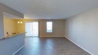 This single-floor residential space offers a clean, functional layout designed for comfortable everyday living. The main living area is a standout feature, showcasing smooth hardwood-style flooring that runs throughout, contributing to a cohesive and modern aesthetic. Neutral beige walls provide a versatile backdrop, allowing the space to feel open and adaptable to a variety of interior styles.

Natural light plays a significant role in the ambiance of the living area, which benefits from a large sliding glass door fitted with vertical blinds, as well as an additional window that together bathe the room in bright, diffused daylight. The sliding door also provides direct access to an outdoor area, enhancing the sense of connection to the exterior.

A textured ceiling adds a subtle layer of character to the space, while clean white baseboards frame the floors with a crisp, finished look. A glimpse of the kitchen area is visible from the main living space, suggesting an open or semi-open floor plan that promotes easy flow between living and cooking areas. The kitchen features a countertop overhang ideal for casual seating. Overall, this space offers a bright, well-maintained environment with practical finishes throughout.