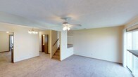 This 2-floor home offers a straightforward and functional layout designed for comfortable everyday living. The main living area is generously sized, featuring wall-to-wall neutral-toned carpet that adds warmth underfoot. A ceiling fan with a built-in light fixture is centrally mounted to the textured ceiling, providing both air circulation and ambient lighting. A chandelier is visible in an adjacent area, hinting at a defined dining or entryway space nearby.

The staircase, finished with carpeted treads and accompanied by a white railing, provides access to the second floor and is neatly integrated into the layout without disrupting the open feel of the main level. Neutral beige walls throughout create a clean, versatile backdrop. A doorway visible in the background leads to an additional room or hallway, suggesting a practical flow between spaces.

The overall ambiance is calm and understated, with a simple, move-in-ready aesthetic. The absence of bold design elements keeps the space flexible, allowing for a variety of interior styles. The two-floor configuration provides separation between living and private spaces, making this home well-suited for a range of occupants.