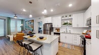Located in the Dr. Phillips area of Orlando, Florida, this single-floor residence offers a thoughtfully designed open-concept layout with modern finishes throughout. The kitchen is a standout feature, showcasing crisp white cabinetry, granite countertops, a center island with bar seating, stainless steel appliances, and pendant lighting — all set atop warm wood-look flooring that flows seamlessly into the living area. The living room is bright and welcoming, with multiple windows dressed in stylish curtains, a ceiling fan, and direct sightlines to the bedrooms. Each bedroom is carpeted for comfort and benefits from natural light through well-appointed windows. The primary bathroom is particularly well-appointed, featuring a backlit vanity mirror, granite countertops, wood-look flooring, and a walk-in closet. A secondary bathroom offers a full tub-shower combination with large-format tile surround and matching granite vanity. Throughout the residence, neutral wall tones and clean architectural lines create a contemporary, cohesive ambiance, complemented by vibrant artwork that adds energy and personality to each room.