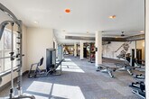 Located on District Lane in Chattanooga, Tennessee, this well-appointed fitness center offers an expansive, single-floor layout designed to accommodate a full spectrum of workout needs. The open floor plan is anchored by structural columns that define distinct exercise zones without compromising the generous sense of space throughout.

The cardio area features a dedicated row of treadmills, ellipticals, and stationary bikes positioned along a windowed wall, flooding the space with natural light and offering views of the surrounding landscape. Ceiling fans overhead ensure comfortable air circulation during intense workouts.

The strength training section is comprehensively equipped with a variety of cable machines, multi-functional weight stations, adjustable benches, and a full dumbbell rack. Exposed industrial-style ductwork runs along the ceiling, lending the space a modern, urban aesthetic. Wall-mounted mirrors amplify the sense of space and allow users to monitor their form.

A distinctive artistic mural featuring silhouetted figures in motion adorns one wall, adding a dynamic visual element that reinforces the fitness-focused atmosphere. The patterned carpet flooring with yellow accent lines provides both durability and a contemporary design touch. Warm, neutral-toned walls and strategically placed recessed and accent lighting create an energizing yet comfortable environment suited to all levels of fitness enthusiasts.