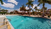 Located at 100 Treviso Grand Cir in Venice, Florida, this outdoor amenity space embodies the relaxed elegance of Southwest Florida resort living. The centerpiece is a generously sized resort-style swimming pool with a shallow entry, sparkling blue water, and an expansive pool deck lined with modern lounge chairs and shaded by tall, swaying palm trees. Colorful umbrellas provide additional shade, creating an inviting atmosphere for leisure and relaxation under the Florida sun.

Surrounding the pool, meticulously maintained tropical landscaping features a variety of palms, shade trees, ornamental shrubs, and flowering plants, all woven together by winding brick-paver and concrete walkways. A wooden pergola serves as a shaded gathering area, complemented by seating that encourages outdoor socializing. The grounds also include a tranquil pond with scenic waterfront views, adding a serene natural element to the community setting.

Curving pedestrian paths meander throughout the property, bordered by lush greenery and mature trees, offering a pleasant environment for walking. The architectural backdrop features Mediterranean-inspired buildings with red-tile roofs, reinforcing the community's cohesive aesthetic. The entire outdoor space is thoughtfully designed to balance active recreation with peaceful natural surroundings.