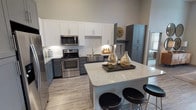 This thoughtfully designed single-floor residence offers a seamless blend of modern finishes and functional living spaces. The kitchen is a standout feature, showcasing two-tone cabinetry with white upper cabinets and slate-gray lower cabinets, granite countertops, a large center island with bar seating, white subway tile backsplash, and a full suite of stainless steel appliances including a refrigerator, range, and built-in microwave. Wood-look flooring flows throughout the main areas, adding warmth and continuity.

The living area is bathed in natural light through tall floor-to-ceiling windows and French doors, creating an open, airy ambiance with generous ceiling height. The bedroom is comfortably sized, featuring large windows, plush carpeting, and a ceiling fan, while a dedicated walk-in closet offers built-in shelving and hanging rods for ample storage.

The primary bathroom is elegantly appointed with a double-sink vanity, granite countertops, white cabinetry, a large mirror, and wood-look flooring. A separate tub and shower area adds convenience. A second walk-in closet with wrap-around built-in organizers provides exceptional storage capacity. The overall aesthetic is contemporary and cohesive, with a neutral gray palette accented by modern fixtures throughout.
