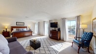This single-floor space centers around a generously proportioned primary bedroom suite designed for comfort and functionality. The bedroom itself is bathed in natural light streaming through multiple large windows and a sliding glass door that opens to an outdoor deck, offering pleasant views of the surrounding landscape. Vaulted ceilings lend an airy, open quality to the room, which is finished with soft wall-to-wall carpeting and a textured ceiling. A patterned area rug anchors the space, complementing the warm tones of the rich wood furniture throughout.

A dedicated sitting area within the suite provides a comfortable retreat, furnished with a sofa, an upholstered accent chair, and a classic dresser with mirror — creating a relaxed lounge space separate from the sleeping area. The bedroom also features a wall-mounted television and an additional chest of drawers, offering ample storage.

The en-suite bathroom is finished in a clean, neutral palette with white cabinetry, a wide vanity with a large mirror, bright overhead lighting, a soaking tub, a separate shower, and a tall linen tower for added storage. Together, these spaces form a cohesive, well-appointed suite that balances practicality with a calm, residential ambiance.