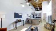This single-floor loft showcases a compelling blend of industrial and contemporary design. Soaring ceilings with exposed wooden beams and visible ductwork create a striking overhead aesthetic, while a skylight and ceiling fan add both natural light and practical comfort to the living area. The open-concept layout seamlessly connects the living and kitchen spaces, fostering a sense of spaciousness throughout.

The kitchen is a standout feature, equipped with stainless steel appliances including a refrigerator, microwave, and gas range. Wood-toned cabinetry pairs elegantly with granite countertops, and a kitchen island with bar-height metal stools serves as both a functional prep area and a casual dining spot. Pendant lighting illuminates the workspace with a warm industrial glow.

The living area is thoughtfully appointed with a wall-mounted flat-screen television, a sleek console table, and a coffee table that anchor the space. Polished concrete floors run throughout, reinforcing the loft's urban character while remaining easy to maintain. The combination of white walls and raw architectural elements creates a balanced, modern atmosphere that is both visually dynamic and inviting.