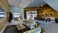 This thoughtfully designed single-floor space makes a striking impression with its dramatic vaulted and coffered ceilings, which lend a sense of grandeur and openness to the interior. The main lounge area is anchored by a plush, cream-toned sectional sofa positioned over a richly patterned area rug, complemented by a rustic wood coffee table and coordinating accent furnishings. A vibrant color palette of teal, mustard yellow, and warm brown ties the space together cohesively.

Large floor-to-ceiling glass doors and expansive windows flood the interior with natural light and offer views of an outdoor patio area beyond, seamlessly connecting the indoors with the exterior. Track and pendant lighting throughout adds warmth and dimension to the space in the evenings.

Adjacent to the lounge, a bar-height counter with upholstered seating provides a social gathering spot, while a fully equipped kitchen with stainless steel appliances is visible in the background, suggesting the space is well-suited for events and entertaining. The open-concept layout encourages easy flow between the lounge, dining, and kitchen zones, making this a versatile and welcoming environment for a variety of gatherings.