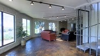 This single-floor space makes a strong architectural impression through its combination of rugged natural materials and thoughtful design. The main living area is anchored by striking stone-clad walls that lend a warm, earthy character to the interior. A wood-burning fireplace serves as a focal point, framed by the same stonework that defines the room's aesthetic. Underfoot, large hexagonal terracotta tiles extend throughout the space, adding texture and a warm, rustic tone to the floor.

The room is generously proportioned, offering ample space for multiple seating arrangements. Leather sofas and accent chairs are arranged to encourage conversation and relaxation, while a central coffee table grounds the grouping. Track lighting runs along the ceiling, providing adjustable, even illumination across the living area, complementing the textured stucco ceiling above.

Large windows line the walls, flooding the interior with natural light and offering leafy, green outdoor views that bring the surrounding landscape into the room. The combination of natural stone, warm tile, and abundant greenery visible through the windows creates a grounded, inviting ambiance. The overall atmosphere is one of comfortable, casual sophistication with a distinctly organic character throughout.