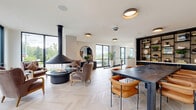 Located in Woodinville, Washington, this thoughtfully designed single-floor commercial amenity space offers a generous footprint ideal for both community gathering and focused work. The layout transitions seamlessly between distinct functional zones, each finished with careful attention to detail and material quality.

The centerpiece of the main lounge area is a striking suspended black fireplace, anchored by a circular seating arrangement of warm leather and velvet chairs. Expansive floor-to-ceiling windows on multiple sides flood the space with natural light and frame lush Pacific Northwest greenery. Underfoot, a light-toned herringbone wood floor adds a refined, geometric elegance throughout this open gathering area.

An adjacent dining and meeting zone features a large dark-topped table with cognac leather seating and a dramatic built-in shelving unit with dark cabinetry, open shelves, and curated décor. A hallway corridor connects to more intimate areas, including a private workstation alcove finished in deep charcoal tones with a built-in desk, decorative stone-look wall tile, and a frosted glass sliding door for privacy.

The carpeted lounge and reception wing provides a softer, more transitional environment with curved contemporary seating, potted greenery, and views of the surrounding landscape. Throughout the space, a cohesive palette of warm neutrals, dark accents, and natural materials creates a polished, welcoming atmosphere.