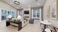 Located at 1352 Town Square Dr in Kent, Ohio, this thoughtfully designed single-floor community space offers a welcoming and refined atmosphere across its well-appointed interior. The main living area is the centerpiece of the space, showcasing soaring vaulted ceilings, light hardwood flooring, and a dramatic floor-to-ceiling stacked-stone fireplace with a built-in linear electric hearth and wall-mounted television above. A statement ring chandelier with warm brass accents anchors the seating area, which is bathed in natural light from multiple large windows dressed in flowing white curtains that offer views of the surrounding outdoor landscape. Recessed lighting throughout enhances the bright, airy ambiance. A cozy nook with a bistro-style table and seating sits beneath vaulted ceilings, providing a relaxed retreat within the space. The kitchenette is outfitted with rich dark-stained wood cabinetry, a marble-look quartz countertop island with bar seating, stainless steel appliances, tile backsplash, and open floating shelves for added functionality. French doors connect the interior to a patio, extending the usable space outdoors. The overall design blends contemporary and transitional elements for a polished, inviting environment.