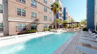 This residential community showcases two well-appointed outdoor pool areas, each reflecting a distinct architectural character. The first pool area is set against a modern multi-story building clad in warm brick and contrasting blue-gray siding. This sparkling pool features built-in water features along its edge, creating a resort-like atmosphere. A pergola-covered seating area with umbrella tables provides shaded outdoor dining nearby, while mature palm trees add a subtropical ambiance under open skies.

The second pool area is framed by a more traditional brick building with pitched rooflines, arched windows, and stucco accents. Curvilinear pool edges are surrounded by a spacious brick-paved deck furnished with blue chaise lounges and umbrella-shaded tables, ideal for relaxing outdoors. Mature trees provide natural shade throughout the courtyard, while manicured shrubs and well-maintained lawn areas enhance the greenery. Private balconies overlooking the pool area are visible on upper floors, offering residents elevated outdoor vistas.

Throughout both areas, thoughtful landscaping with layered plantings, ground cover, and ornamental trees creates a cohesive, park-like setting. The overall ambiance is inviting and well-maintained, blending classic architectural details with comfortable, resort-inspired outdoor amenities.