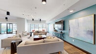 Located at 355 N Rosalind Ave in the heart of Orlando, FL, this generously proportioned single-floor amenity space offers a thoughtfully designed communal environment spanning over 3,000 square feet. The open-concept layout is defined by wide-plank wood-look flooring, crisp white walls, and strategically placed structural columns that subtly delineate distinct activity zones without compromising the sense of openness.

The main lounge area features multiple seating arrangements, including soft neutral sectionals anchored by a teal accent wall with a large mounted flat-screen television and curated artwork. A separate conversation zone showcases teal rounded accent chairs arranged around a central coffee table beneath a striking cross-shaped linear light fixture. Additional seating nooks with vibrant red accent chairs are positioned near floor-to-ceiling windows that flood the space with natural light and offer views of the surrounding streetscape.

A bar-height counter with wood stools serves as a casual dining or social area, while bicycle-themed wall art and gallery arrangements lend the space a dynamic, urban character. A wide corridor with a circular ceiling fixture and decorative credenza connects the zones seamlessly. Glass doors open directly onto an outdoor terrace furnished with additional seating. The overall ambiance is modern, bright, and inviting, suited for both social gatherings and relaxed everyday use.