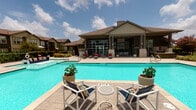 Located at 5702 Timbergate Dr in Corpus Christi, Texas, this single-story community clubhouse and amenity center is designed to deliver a resort-like experience. The exterior showcases a distinctive combination of stone masonry columns, board-and-batten green siding, and a pitched shingle roof, lending the building a polished yet welcoming character.

At the heart of the outdoor amenities is a generously sized swimming pool with clear turquoise water, surrounded by a spacious stamped concrete deck furnished with colorful lounge chairs and potted plants. The pool area is enclosed by a classic wrought-iron fence with stone pillar accents, providing both security and aesthetic appeal, with swaying palm trees visible beyond the perimeter.

A covered outdoor lounge area serves as a natural gathering space, anchored by a striking floor-to-ceiling stone fireplace topped with a wall-mounted flat-screen television. Comfortable seating with bold navy and multicolored cushions creates an inviting atmosphere for relaxation. Adjacent to the lounge, a covered patio with outdoor dining furniture offers additional seating, framed by expansive floor-to-ceiling glass walls that connect seamlessly to the interior and provide unobstructed views of the pool. The abundant natural light and open-air design make this amenity space ideal for both social gatherings and quiet enjoyment year-round.
