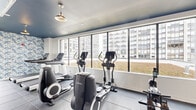 This fitness facility is thoughtfully designed to support a comprehensive workout experience across multiple distinct zones. The space is unified by dark slate tile flooring and a striking navy ceiling, with exposed industrial-style ductwork adding an urban, modern character throughout.

The cardio zone is lined with large floor-to-ceiling windows that frame views of the surrounding city skyline, flooding the area with natural light. It features a full lineup of Life Fitness equipment, including treadmills, ellipticals, and stationary bikes, all oriented toward the expansive windows for an energizing workout environment.

A separate functional training area is distinguished by vibrant blue-and-white tropical leaf wallpaper, adding a bold visual contrast to the space. This zone includes a cable machine, a full rack of dumbbells, kettlebells, medicine balls, barbells, and a large wall mirror for form-checking during strength sessions. A utility counter and additional amenities are conveniently situated nearby.

The facility also includes access to an outdoor terrace surfaced with artificial turf, accented by natural wood plank planters and framed by metal railings. The terrace offers open views of the urban skyline, providing an inviting space for outdoor activity or relaxation. Together, the indoor and outdoor areas create a versatile and well-appointed fitness destination.