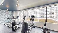 This fitness facility is thoughtfully designed to support a comprehensive workout experience across multiple distinct zones. The space is unified by dark slate tile flooring and a striking navy ceiling, with exposed industrial-style ductwork adding an urban, modern character throughout.

The cardio zone is lined with large floor-to-ceiling windows that frame views of the surrounding city skyline, flooding the area with natural light. It features a full lineup of Life Fitness equipment, including treadmills, ellipticals, and stationary bikes, all oriented toward the expansive windows for an energizing workout environment.

A separate functional training area is distinguished by vibrant blue-and-white tropical leaf wallpaper, adding a bold visual contrast to the space. This zone includes a cable machine, a full rack of dumbbells, kettlebells, medicine balls, barbells, and a large wall mirror for form-checking during strength sessions. A utility counter and additional amenities are conveniently situated nearby.

The facility also includes access to an outdoor terrace surfaced with artificial turf, accented by natural wood plank planters and framed by metal railings. The terrace offers open views of the urban skyline, providing an inviting space for outdoor activity or relaxation. Together, the indoor and outdoor areas create a versatile and well-appointed fitness destination.