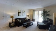 This single-floor space offers a comfortable and thoughtfully designed living environment. The living room serves as the centerpiece of the home, featuring plush carpeting underfoot and a neutral color palette that creates a warm, inviting atmosphere. Large sliding glass doors flood the room with natural light while providing views of the outdoors and access to a balcony or patio area, adding to the overall sense of openness. The living area is anchored by a dark upholstered sofa accented with patterned throw pillows, paired with a dark wood coffee table and coordinating side tables, creating a cohesive and stylish aesthetic. Elegant floor lamps and table lamps provide warm ambient lighting throughout the space. Decorative wall art adds a modern touch to the light-painted walls, while a tall indoor plant in the corner brings a refreshing natural element to the interior. The ceiling features a clean, paneled finish that complements the overall design. The space strikes a balance between functionality and style, making it well-suited for everyday living and relaxed entertaining. The abundant natural light and access to the outdoors enhance the bright, airy feel of the interior.