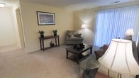 This single-floor residence offers a clean, comfortable living environment designed with functionality and ease in mind. The living room serves as the heart of the space, showcasing wall-to-wall carpeting that adds warmth underfoot and a soft, neutral color palette across the walls that creates a calm, inviting atmosphere. A large window dressed with vertical blinds allows generous natural light to filter in, brightening the room throughout the day. The space is thoughtfully laid out to accommodate relaxed seating arrangements, with ample room for a sofa, accent chairs, and a central coffee table. A console table along one wall provides additional surface space and display area, complemented by decorative greenery that adds a touch of life to the room. Framed wall art contributes a finishing touch to the overall aesthetic. The combination of soft lighting from a floor lamp and the natural light from the window creates a balanced and pleasant ambiance. The overall design of the space leans toward a practical yet comfortable style, making it well-suited for everyday living. The single-floor layout ensures easy accessibility and a seamless flow throughout the residence.