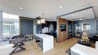 The clubhouse at 1810 State Street in San Diego is a single-story amenity space designed with a sophisticated, contemporary aesthetic. The open-concept layout integrates a fully equipped kitchen, lounge seating, and entertainment areas into one cohesive environment, making it well-suited for both casual gatherings and organized events.

The kitchen serves as a central focal point, anchored by a white quartz island with seating for multiple guests along gold-accented bar stools. Dark cabinetry, stainless steel appliances, a white subway tile backsplash, and a pendant light fixture with a geometric brass frame contribute to the upscale, curated design. Light wood-toned flooring runs throughout, providing a warm contrast to the deep navy walls trimmed in gold detailing.

A warm walnut wood accent wall frames a wall-mounted flat-screen television, creating a dedicated media area that transitions seamlessly into the lounge zone. Recessed lighting throughout ensures even, ambient illumination. Additional seating and a gaming area are visible, offering multiple activity zones within the space. The overall ambiance balances bold design choices with functional versatility.