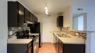 Located at 5400 Diamond Loch in Columbus, Ohio, this single-floor unit offers a practical and well-appointed living space with a cohesive design throughout. The open-concept living area is finished with neutral wall-to-wall carpeting, a textured ceiling, and a ceiling fan with integrated lighting, creating a comfortable and airy atmosphere. A half-wall partition separates the living area from the kitchen, maintaining an open sightline while defining each space. The kitchen is a standout feature, designed as a galley-style layout with dark espresso cabinetry that contrasts sharply against light countertops. A mosaic tile backsplash in earth tones adds a stylish, contemporary touch. The kitchen is equipped with black and stainless steel appliances, including a range, refrigerator, and overhead microwave. A stainless steel sink is positioned beneath upper cabinetry, and pendant lighting overhead adds a modern accent. Light hardwood-style flooring runs throughout the kitchen, complementing the overall aesthetic. The space combines functional design with updated finishes, making it a well-suited home for everyday living in Columbus's west side.