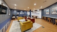 Located at 201 Munson Street in New Haven, CT, this single-floor amenity space serves as a dynamic recreation and social lounge. The room is defined by a bold navy and white color scheme, with deep blue accent walls contrasting against crisp white surfaces and warm light wood flooring throughout. Recessed lighting illuminates the space evenly, creating a bright and energetic atmosphere.

A prominent "Work Hard Play Harder" mural anchors the game zone, which is equipped with a foosball table, a shuffleboard table, and arcade-style machines — offering ample entertainment options. Framed artwork and decorative pieces adorn the walls, adding a curated, residential touch to the space.

The lounge area is thoughtfully furnished with a rich cognac leather sofa, mustard yellow accent chairs, and a marble-top coffee table, all arranged atop a layered area rug. A wall-mounted flat-screen TV faces the seating arrangement, making the space equally suited for relaxed viewing. A built-in counter with bar-height seating along one wall provides a dedicated workspace or casual dining area. Structural columns are finished in coordinating navy tones, seamlessly integrating into the overall design. The space balances energetic recreation with comfortable lounging in a cohesive, well-appointed environment.