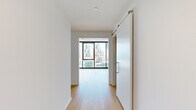 This thoughtfully designed unit accommodates three bedrooms, one bathroom, a combined kitchen and living area, and two hallways across a single floor. The architectural character is defined by exposed concrete ceilings paired with crisp white walls and light-toned hardwood flooring, creating a clean industrial-modern aesthetic throughout.

The living area benefits from floor-to-ceiling windows framed in black metal, offering expansive city skyline views and abundant natural light. A sliding barn-style door connects the living space to the hallway, adding a modern functional detail. Track lighting complements the concrete ceiling in the main living zone.

The kitchen features light wood cabinetry, white lower cabinets, a neutral countertop, textured tile backsplash, a matte black faucet with stainless steel sink, and stainless steel appliances including a refrigerator, dishwasher, oven, and built-in microwave.

The bedrooms continue the floor-to-ceiling window theme, maximizing city views and natural light, with exposed concrete ceilings lending an urban character. One bedroom includes a built-in shelved walk-in closet.

The bathroom features a floating double-vanity with dark wood cabinetry, dual undermount sinks, matte black fixtures, rounded rectangular mirrors, and a glass-enclosed shower.