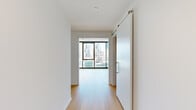 This thoughtfully designed unit accommodates three bedrooms, one bathroom, a combined kitchen and living area, and two hallways across a single floor. The architectural character is defined by exposed concrete ceilings paired with crisp white walls and light-toned hardwood flooring, creating a clean industrial-modern aesthetic throughout.

The living area benefits from floor-to-ceiling windows framed in black metal, offering expansive city skyline views and abundant natural light. A sliding barn-style door connects the living space to the hallway, adding a modern functional detail. Track lighting complements the concrete ceiling in the main living zone.

The kitchen features light wood cabinetry, white lower cabinets, a neutral countertop, textured tile backsplash, a matte black faucet with stainless steel sink, and stainless steel appliances including a refrigerator, dishwasher, oven, and built-in microwave.

The bedrooms continue the floor-to-ceiling window theme, maximizing city views and natural light, with exposed concrete ceilings lending an urban character. One bedroom includes a built-in shelved walk-in closet.

The bathroom features a floating double-vanity with dark wood cabinetry, dual undermount sinks, matte black fixtures, rounded rectangular mirrors, and a glass-enclosed shower.