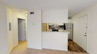 This one-bedroom, one-bathroom residence is thoughtfully laid out across a single floor, offering a functional and comfortable living environment. The open living area features plush wall-to-wall carpeting, neutral tones, and a ceiling fan with light fixture overhead. A large window and a sliding glass door flood the space with natural light while providing direct access to a private balcony with outdoor views.

The kitchen is designed around a peninsula-style counter with a light speckled surface, creating a natural division between the cooking and living areas. White cabinetry, a stone tile backsplash, and stainless steel appliances—including a range and refrigerator—give the kitchen a clean, practical aesthetic. Wood-look vinyl flooring runs throughout the kitchen and entry areas, offering a durable contrast to the carpeted living and sleeping spaces.

The bedroom continues the carpeted flooring and neutral palette, with a sliding glass door providing additional natural light and balcony access. The bathroom includes a full soaking tub and shower combination with white tile surround and chrome fixtures. A dedicated utility area accommodates a water heater, adding to the unit's self-contained functionality.