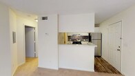 This one-bedroom, one-bathroom residence is thoughtfully laid out across a single floor, offering a functional and comfortable living environment. The open living area features plush wall-to-wall carpeting, neutral tones, and a ceiling fan with light fixture overhead. A large window and a sliding glass door flood the space with natural light while providing direct access to a private balcony with outdoor views.

The kitchen is designed around a peninsula-style counter with a light speckled surface, creating a natural division between the cooking and living areas. White cabinetry, a stone tile backsplash, and stainless steel appliances—including a range and refrigerator—give the kitchen a clean, practical aesthetic. Wood-look vinyl flooring runs throughout the kitchen and entry areas, offering a durable contrast to the carpeted living and sleeping spaces.

The bedroom continues the carpeted flooring and neutral palette, with a sliding glass door providing additional natural light and balcony access. The bathroom includes a full soaking tub and shower combination with white tile surround and chrome fixtures. A dedicated utility area accommodates a water heater, adding to the unit's self-contained functionality.