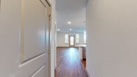 This single-floor unit showcases a clean, contemporary design characterized by neutral tones, recessed lighting, and rich hardwood-style flooring that flows consistently throughout the space. The open-concept layout connects the living area and kitchen seamlessly, creating an airy and functional environment.

The kitchen is a standout feature, outfitted with white shaker-style cabinetry, a full suite of stainless steel appliances including a refrigerator, range, microwave, and dishwasher, and a central island with a sink and light quartz countertop. Globe pendant lights hang above the island, adding a modern aesthetic touch.

The living area is generously proportioned, bathed in natural light from large windows flanking a rear entry door. The high ceilings enhance the open feel of the space, while recessed lighting provides even illumination throughout.

The bedroom features dual windows for natural light, a ceiling fan, and ample space. The bathroom is finished with dark subway tile surrounding the tub-shower combination, complemented by hardwood-style flooring and clean white fixtures. A built-in desk nook provides a practical workspace within the unit.