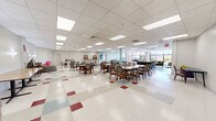 Located at 860 E Broadway in Long Beach, NY, this expansive single-floor community facility offers a generous, open footprint designed to accommodate a variety of activities and gatherings. The interior features a classic drop-panel ceiling with recessed fluorescent lighting, and a distinctive terrazzo-style floor with colorful tile accents that adds character throughout the space.

The main hall serves as a versatile dining and activity area, furnished with multiple rectangular and round tables paired with padded chairs, accommodating both intimate gatherings and larger group events. Stacked folding tables and round tabletops indicate the room's adaptability for varied configurations. A structural column anchors the center of the room, while accent walls in soft aqua tones provide visual interest and a welcoming ambiance.

A lounge area near the floor-to-ceiling glass window wall offers comfortable seating with sofas arranged around a wall-mounted flat-screen TV, bathed in natural light and offering views of an outdoor patio space. Adjacent to this, a fitness area is equipped with stationary bikes and exercise equipment, also benefiting from the same expansive glazed facade that connects the interior to the outdoors.

Additional spaces include a quieter anteroom with a podium and seating, suitable for presentations or private meetings. An American flag on display adds a sense of community pride to this well-maintained, functional facility.