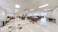 Located at 860 E Broadway in Long Beach, NY, this expansive single-floor community facility offers a generous, open footprint designed to accommodate a variety of activities and gatherings. The interior features a classic drop-panel ceiling with recessed fluorescent lighting, and a distinctive terrazzo-style floor with colorful tile accents that adds character throughout the space.

The main hall serves as a versatile dining and activity area, furnished with multiple rectangular and round tables paired with padded chairs, accommodating both intimate gatherings and larger group events. Stacked folding tables and round tabletops indicate the room's adaptability for varied configurations. A structural column anchors the center of the room, while accent walls in soft aqua tones provide visual interest and a welcoming ambiance.

A lounge area near the floor-to-ceiling glass window wall offers comfortable seating with sofas arranged around a wall-mounted flat-screen TV, bathed in natural light and offering views of an outdoor patio space. Adjacent to this, a fitness area is equipped with stationary bikes and exercise equipment, also benefiting from the same expansive glazed facade that connects the interior to the outdoors.

Additional spaces include a quieter anteroom with a podium and seating, suitable for presentations or private meetings. An American flag on display adds a sense of community pride to this well-maintained, functional facility.