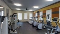 This fitness center is designed to support a well-rounded workout routine within a comfortable, single-floor space. The room is outfitted with multiple Precor cardio machines, including treadmills and ellipticals, arranged to allow users ample space to exercise without feeling cramped. A strength training station is positioned along the left wall, providing options for resistance and weight-based workouts. The flooring consists of durable rubber matting, a practical choice that provides cushioning and stability during exercise. Natural light filters into the room through several windows fitted with white horizontal blinds, contributing to an open and energizing atmosphere. Overhead, flush-mount light fixtures and a ceiling fan ensure the space remains well-lit and properly ventilated. The walls feature a neutral beige tone accented by bold horizontal stripes of orange and gray, adding a dynamic visual element to the room's design. A wall-mounted television is positioned for visibility during cardio sessions. The overall ambiance is clean, functional, and purposefully arranged to serve as a practical fitness facility.