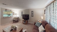 This single-floor residence offers a well-organized and comfortable layout across several rooms, each finished with warm neutral tones and plush carpeting throughout. The living room serves as the central gathering space, featuring crown molding, a textured ceiling, and ample room for seating and entertainment. Decorative wall accents and a large window with patterned curtains add character and allow natural light to fill the space.

The kitchen is efficiently designed with honey-toned wood cabinetry, laminate countertops, a double-basin sink, and white appliances including a coil-burner range and refrigerator. A range hood is centrally positioned above the stove, and the layout makes practical use of available space.

The home includes multiple bedrooms, each equipped with ceiling fans and textured ceilings. The primary bedroom features a sliding closet, a window with sheer curtains for soft natural light, and space for a dresser and media setup. A second bedroom offers a cozy retreat with darker curtains and a ceiling fan with lighting. A third, more compact bedroom provides additional sleeping space with a textured ceiling and neutral walls.

The bathroom includes a single-sink vanity with wood cabinetry, a large mirror, and wall-mounted lighting, offering a clean and functional layout. Overall, this home presents a practical and cohesive living environment.