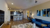 This single-floor residence showcases an open-concept design that seamlessly integrates the kitchen, dining, and living areas into one cohesive, well-lit space. The kitchen is outfitted with dark espresso cabinetry, stainless steel appliances including a refrigerator, stove, and microwave, and a central island with bar seating for three. Pendant lighting hangs above the island, adding a warm, focused glow to the prep area, while recessed ceiling lights illuminate the broader space. The wood-look flooring extends throughout, providing a consistent and warm visual flow. Adjacent to the kitchen, a dedicated dining area features a dark wood table paired with upholstered seating. The living area is anchored by a bold blue sofa, a geometric-patterned area rug, and a gold-accented coffee table, contributing a contemporary aesthetic. A glimpse down a hallway reveals access to what appears to be a bathroom with tiled flooring and warm lighting. The neutral wall tones throughout complement the darker furnishings and cabinetry, resulting in a balanced and inviting atmosphere.