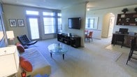 This single-story space is designed with an open-concept layout that encourages a natural flow between the living and dining areas. The living room is anchored by plush wall-to-wall carpeting and features a wall-mounted flat-screen television positioned above a sleek, dark entertainment console. A glass-topped coffee table with slender legs sits at the center of the seating area, adding a contemporary touch. Large windows along the exterior wall are dressed with patterned curtains, allowing generous amounts of natural light to fill the space while maintaining privacy.

A structural column with decorative molding detail subtly divides the living area from a connected dining nook, where a compact dining set with cushioned chairs creates an intimate setting. An arched doorway is visible to one side, adding an architectural accent that softens the overall aesthetic. A large mirror positioned near the dining area reflects additional light, enhancing the sense of openness. The neutral green-toned walls complement the light carpeting throughout, contributing to a calm and cohesive ambiance across the entire space.