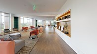 Located at 12 Leo M Birmingham Pkwy in Boston's Brighton neighborhood, this beautifully designed amenity floor offers a sophisticated and inviting environment for residents and guests alike. The space is characterized by rich hardwood flooring, crisp white ceilings with warm cove lighting, and expansive floor-to-ceiling windows that flood the interiors with natural light and frame lush outdoor views.

The main lounge area is thoughtfully arranged with multiple seating zones, including a plush sectional sofa anchored by a dramatic dark textured fireplace wall, creating a cozy yet refined focal point. Café-style tables with contemporary chairs are arranged throughout, encouraging casual gatherings and social interaction.

A wide bank of glass folding doors opens directly onto a spacious outdoor terrace, seamlessly blending indoor and outdoor living. The terrace, finished with a pergola structure and additional seating, extends the entertaining potential of the floor considerably.

An extended corridor lounge features built-in wood shelving with warm accent lighting, curated décor, and comfortable seating clusters set against floor-to-ceiling windows overlooking the treetops.

The gourmet kitchen stands out with a striking marble waterfall island, rose gold wire barstools, professional-grade stainless appliances, a statement range hood, and sleek white cabinetry — all bathed in panoramic natural light, making it an ideal space for culinary events and entertaining.