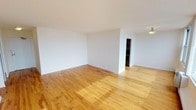 This single-floor space presents a clean, minimalist aesthetic defined by warm-toned hardwood floors and crisp white walls that create a bright, welcoming atmosphere. The main living area is generously proportioned, offering an open feel enhanced by smooth white ceilings and abundant ambient light. The hardwood flooring throughout adds natural warmth and a timeless character to the interior.

A partial dividing wall separates the main area from an adjoining space, providing a degree of architectural definition while maintaining an overall sense of openness and flow. Beyond the partial wall, a connected secondary area is visible, which appears to offer additional functional space such as a closet or utility area, adding to the practical layout of the unit.

The flush ceiling light fixture provides overhead illumination, complementing the natural light that fills the space. The white baseboards and neutral wall tones throughout give the interior a cohesive, well-maintained appearance. The straightforward, uncluttered design of this space makes it highly adaptable, suitable for a variety of uses and furniture arrangements. Overall, the space combines functional simplicity with warm, inviting finishes to create a comfortable and versatile environment.