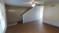 This two-story home offers a straightforward and functional layout spread across two levels connected by an interior staircase. The main living area on the ground floor is finished with warm wood-look laminate flooring that extends throughout the space, providing a cohesive and easy-to-maintain surface underfoot. A ceiling fan with an integrated light fixture is centrally mounted, ensuring comfortable airflow and ambient lighting throughout the room. The staircase, finished with white trim and a wooden handrail, rises along one wall and serves as a visual focal point while efficiently connecting the two floors. The walls are painted in a neutral, light tone that keeps the interior feeling open and bright. A doorway visible from the main living area leads to an adjacent room or corridor, suggesting a well-connected floor plan that allows for natural flow between spaces. The textured ceiling adds a subtle architectural detail consistent throughout the level. Overall, the home presents a clean, move-in ready canvas with practical finishes and a simple, open feel that offers flexibility for a variety of living arrangements across both floors.