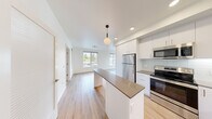 Located at 23040 Military Rd in Kent, WA, this 2-bedroom, 2-bathroom unit offers a well-designed, modern living space across a single floor. The open-concept kitchen and living area serves as the heart of the home, showcasing white shaker cabinetry, dark quartz countertops, a subway tile backsplash, and a full suite of stainless steel appliances including a refrigerator, electric range, and microwave. A central kitchen island with pendant lighting provides additional prep space and adds a stylish focal point that connects seamlessly to the living area, which is bathed in natural light from large windows overlooking the surrounding neighborhood and mature trees. Light wood-style flooring runs throughout the main living areas, complementing the clean, bright aesthetic. The primary bedroom is generously sized, featuring a ceiling fan, plush carpet flooring, and a window with outdoor views, while the second bedroom also offers carpeting and natural light. A spacious walk-in closet with wire shelving provides ample storage. Both bathrooms are finished with white cabinetry, dark countertops, a large mirror, and a tub-shower combination. A bright hallway connects all rooms, maintaining a clean, cohesive flow throughout the unit.