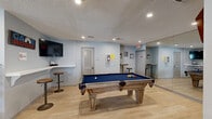 Located in Lee's Summit, Missouri, this single-floor space is a thoughtfully designed combination of a living room and game room, offering a dynamic and entertaining environment. The open layout is unified by light hardwood flooring and a textured ceiling with recessed lighting throughout, creating a bright and welcoming ambiance.

The game room section is a standout feature, anchored by a full-size pool table with a bold blue felt surface and ornate wooden legs. A large wall mirror amplifies the sense of space and light in this area. A dedicated game room nook includes a wall-mounted TV, a built-in bar-height counter with stools, and a compact refrigerator — ideal for entertaining. A dartboard adds to the recreational appeal.

The living area transitions seamlessly from the game room and features a large, tufted leather sectional sofa with colorful accent pillows, centered around a woven ottoman coffee table. Large windows dressed in navy curtains allow natural light to fill the space, while views into an adjacent room add depth. A workstation with a monitor and an arcade game machine round out the multifunctional nature of the lounge area.

The space connects to additional rooms via a hallway, and the overall color palette of soft blues and grays lends a cohesive, modern feel throughout.