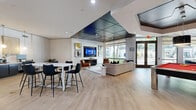This thoughtfully designed single-floor community amenity space in Addison, Texas, offers a generous open-plan layout that seamlessly blends leisure, work, and social zones. Light wood-look flooring flows throughout, providing a warm, cohesive foundation for the varied activity areas within.

A central lounge area anchors the space with a plush neutral sectional sofa, a mid-century leather accent chair, and a mounted flat-screen television, all set beneath a striking dark geometric ceiling panel that adds a bold architectural focal point. Adjacent to the lounge, a red billiards table invites recreation, while a high-top bar-style table with sleek black chairs provides a casual spot for dining or socializing.

A dedicated workstation area features desks and task chairs beneath pendant lighting, ideal for focused work or meetings. Nearby, a café-style nook with rounded tables, vibrant navy blue accent chairs, and botanical wall art sits beside a coffee station framed in dark wood cabinetry.

Floor-to-ceiling windows and glass double doors flood the space with abundant natural light while offering open views of mature trees and landscaped outdoor surroundings. Recessed lighting throughout ensures the space remains bright and inviting at any hour. The overall ambiance is modern, polished, and welcoming — designed to accommodate a wide range of activities in a single, well-appointed venue.
