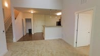 This two-floor residence offers an open and airy layout highlighted by impressively high ceilings that amplify the sense of space throughout the main living area. The living room features neutral carpeting that adds warmth underfoot, while the adjoining kitchen and dining area transition to rich hardwood flooring, creating a natural distinction between spaces. The kitchen is anchored by a generous peninsula with granite countertops, providing both additional prep space and casual seating. Pendant lighting hangs above the peninsula, offering focused illumination and a polished finishing touch. Stainless steel appliances are visible within the kitchen, lending a modern, functional character to the cooking space. The open sightlines from the living area through to the kitchen and beyond create a cohesive, connected atmosphere ideal for entertaining and everyday living. Multiple doorways lead to additional rooms, including what appears to be a closet or utility space, enhancing the overall storage and functionality of the layout. The neutral color palette of warm beige tones throughout the walls and flooring contributes to a bright, welcoming ambiance. Spanning two floors, this residence offers ample room for comfortable living across a well-planned and thoughtfully designed floor plan.